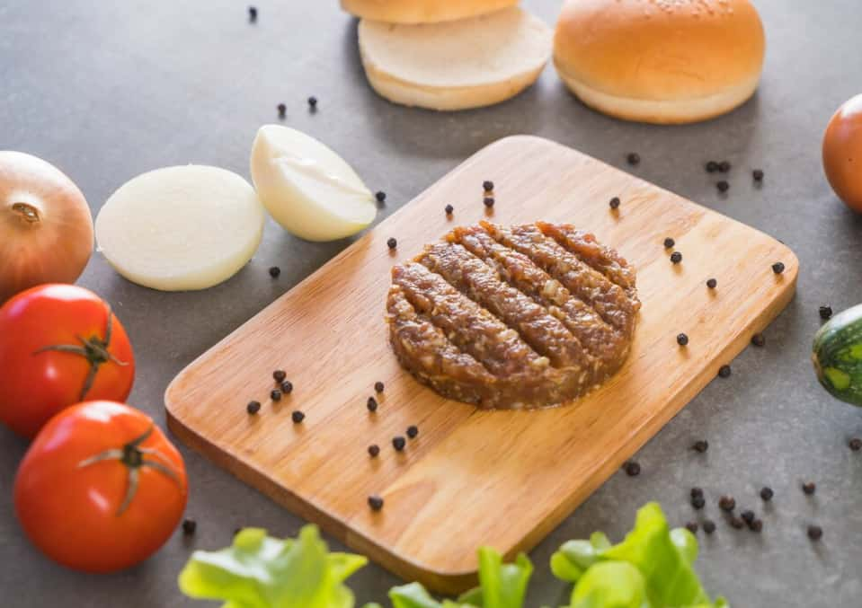 A raw burger patty on a wooden board with ingredients around